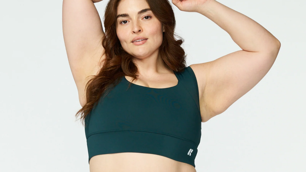 Woman demonstrating open, aligned posture with arms raised in a teal sports bra