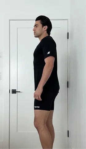A man stands in profile wearing a black t-shirt and shorts, with his shoulders slumped forward.